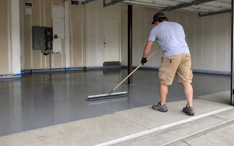 A kneeling applicator presses urethane cement into a coved base along a commercial kitchen wall with a floor drain nearby.