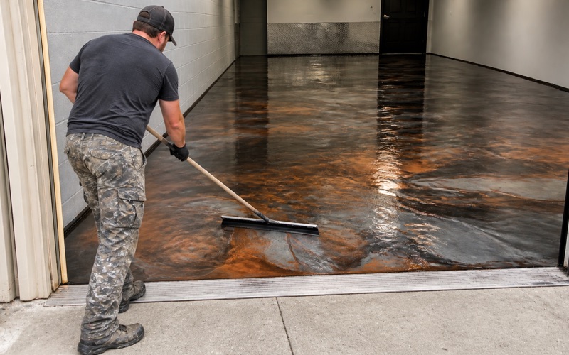 An operator crouches with one hand on the dust shroud collar of a walk-behind grinder that has ground half the shop bay floor open and matte.